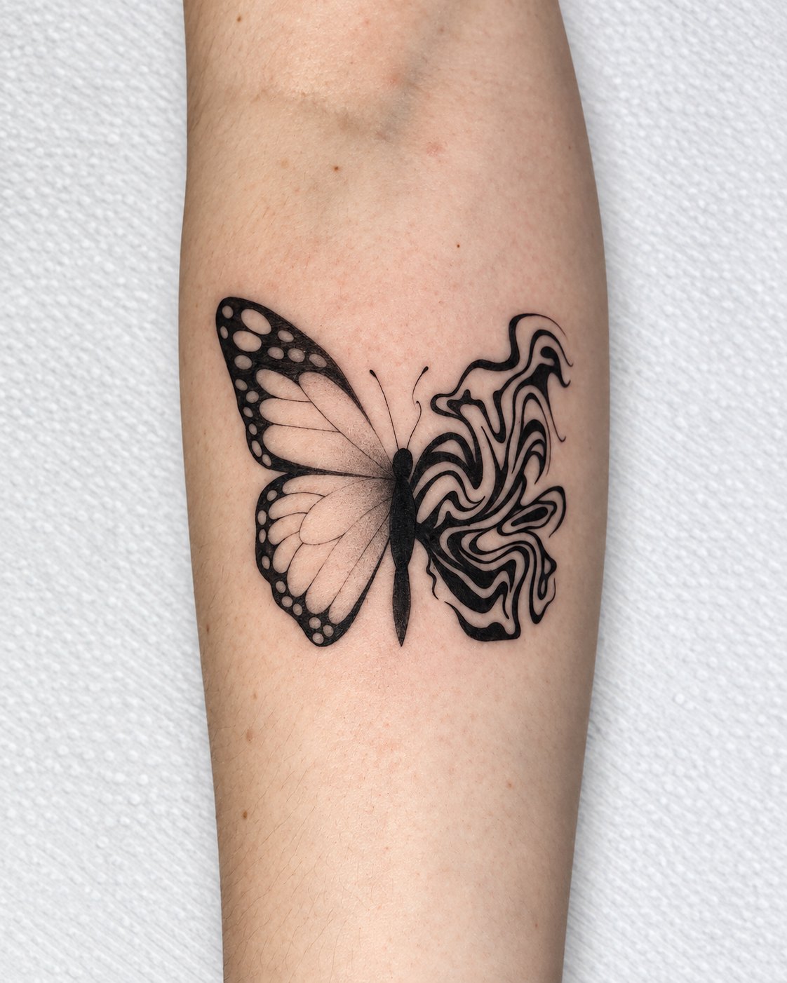 Tatuagem de borboleta no antebraço: uma asa em traço fino com pontilhismo, a outra em ondas de preto sólido